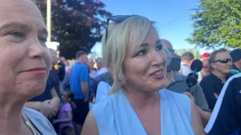 Two smiling ladies, one dark haired and one blond, stand in front of a crowd of people on a bright sunny evening
