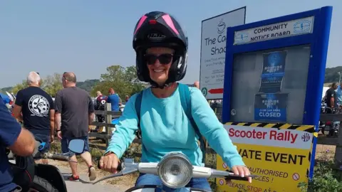 Sandra Baker Ali Richards smiles for the camera as she sits on her white scooter. She has a flip up crash helmet with the face open and a light blue jumper.