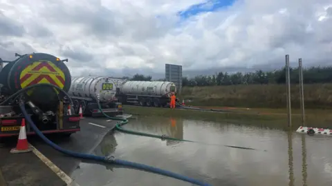 National Highways Three tankers on the A421, clearing water that is on the road surface, with a man standing in an orange florescent suit, by one