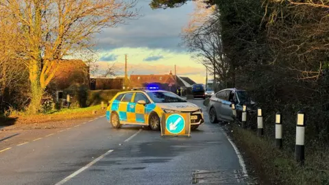 A police car blocking the road. There is a silver car crashed into the trees to the right. 