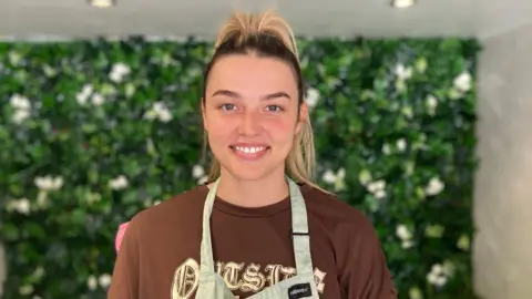 A woman wearing an apron over a burgudy top is smiling directly at the camera. There is a green background behind her