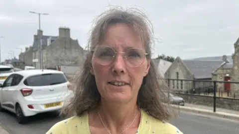 A middle-aged woman with glasses looking at the camera. She is standing beside a road in an urban area. She is wearing a yellow top and the sky is grey.