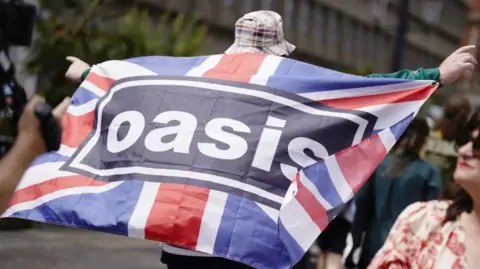 A man is holding a Union Jack flag which has the black and white Oasis logo in the middle. He is wearing a bucket hat and shorts. 