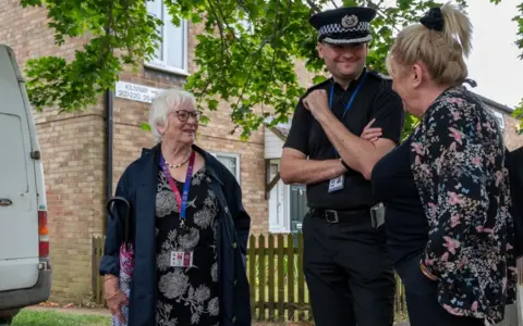 OPFCC A woman in a black floral dress speaks to a police officer and another woman in a blue floral dress in the middle of a housing estate. 