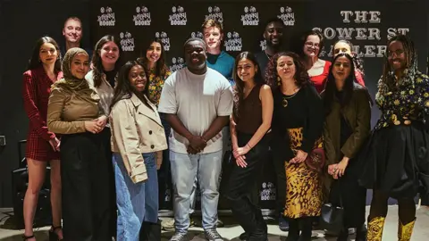 Will Fahy A group of 15 young writers, at an event, with Abaka standing in the middle. The backdrop is a black board with white writing which reads "#Merky Books"