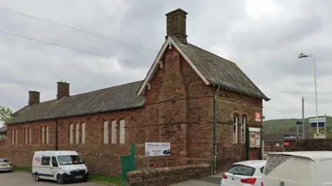 A brick rectangular train station with a gable end and the track just visible behind the main station building 