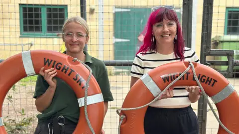 Wightlink A female zoo handler, who has blonde hair and glasses and is wearing a green uniform, stands next to another woman, who has dyed pink hair and is wearing a white top with dark stripes. They are both smiling at the camera and holding up orange life rings. 