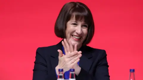 Reuters Chancellor Rachel Reeves wears a black suit jacket and a royal blue shirt as she applauds behind a desk at conference