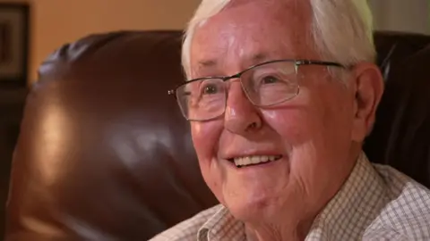 BBC Stephen Harrington is an older man, aged 83. He has white hair and wears thin-rimmed clear-lensed glasses. He is sitting on a brown leather chair and wears a white and brown checked shirt. He is smiling.
