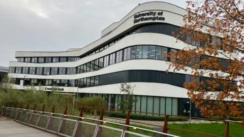 A black and white building surrounded by grass, grey sky and an tree with brown leaves.