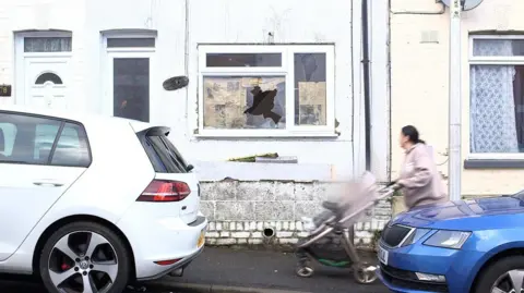 Sarah Brinicombe/BBC Demi Worboys' walking near her temporary accommodation in Chatham, Kent. She is wearing a beige hoodie and is pushing a pram holding her son. A visibly broken window can be seen on the property, which has newspaper around the affected area. 