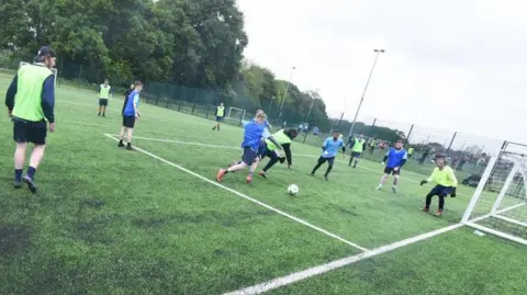 Two football players from rival teams edging closer to the ball as the goal keeper leans forward in anticipation. One team is wearing lime shirts and their opponents are in blue. 