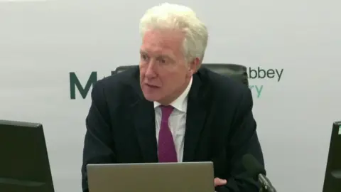 A man sits in front of a laptop and computer monitors. he's wearing a black suit with a white shirt and a pink tie. A mic is in front of him and a backdrop behind him says Muckamore Abbey Hospital Inquiry.