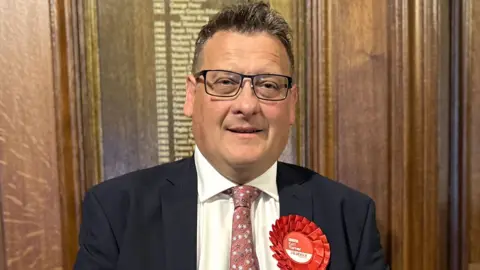 BBC Karl Turner the succesful Labour candidate for Hull East pictured on election night in 2024 wearing a dark suit jacket, white shirt, and patterned pink tie, standing indoors against a wooden paneled wall. A large red rosette with the word ‘Labour’ and smaller text is pinned to the left side of the jacket. 