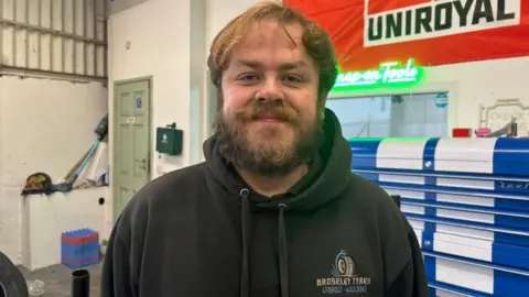 BBC/ Alex James A man in a black hoodie is stood in a car garage. His hoodie has a blue and white logo on the front. On the right are blue and white cupboards above which is a red flag. 