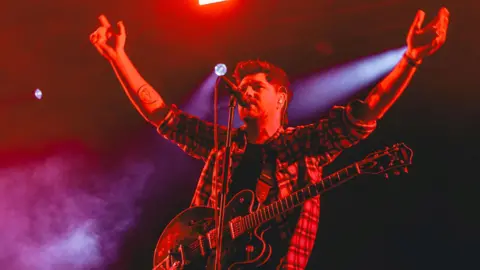 Getty Images Twin Atlantic frontman Sam McTrusty, pictured at the Y Not Festival in 2023, is standing on a stage with a guitar slung over his shoulders and his arms raised. The musician is bathed in a red glow.