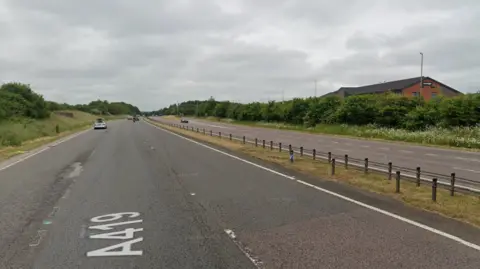 A view looking down the A419 from google maps where is is three lanes. There are a few cars on the road and it is a grey cloudy day. Green verges with the top of a red brick building seen on the right behind them.