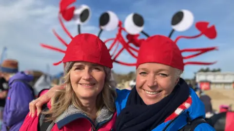 Two women are standing close together wearing bight red hats on a beach. They are smiling and there are other people waking in the background.