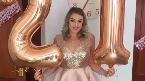 Bethan holding two balloons in the shape of a 21. She is wearing a dress with a sparkly top and a light pink skirt, and is wearing pink lipstick. Her hair is curled. There are pink birthday banners on two doors either side of her.