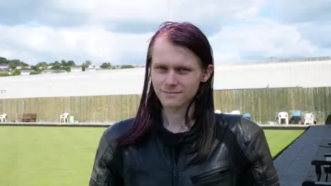 Rowan has long shoulder length purple hair and lighteyebrows. He's wearing a dark leather jacket and is stood on a bowling green