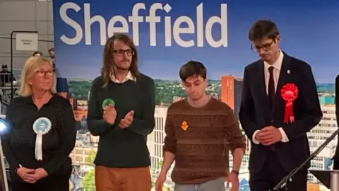 BBC/Simon Thake A woman (far left) and three men are standing in a row at an election count. Each person is wearing a different colour rosette. They are awaiting the outcome of an election count. Andy Davies, wearing beige trousers and a jumper stands second from the left. Tom Hunt, wearing a dark suit and sporting a large red rosette, stands with his hands clasped and his head down on the far right. 