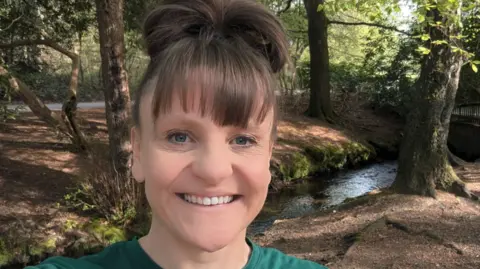 Tonia Dunn A woman with brown hair in a green top is smiling at the camera in Sutton Park. A stream is behind her on the right and trees are also in the background. 