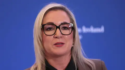 PA Media Michelle O'Neill. She has shoulder-length blonde hair, wearing glasses and a brown blazer. The is a blue background.