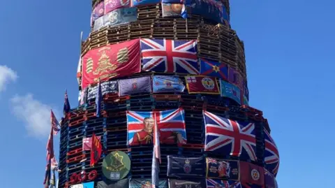 A large number of flags including union flags and a flag depicting an image of King Charles placed on a bonfire in Derry's Bogside.