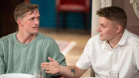 BBC/Studio Lambert/Euan Cherry Two men sitting next to each other. The one on the right is talking across the other, to people who are sat out of frame. The man talking has short brown hair and wears a short-sleeved plain white shirt and is gesticulating. The man on the left looks at him and appears to be listening intently. He wears a light teal knitted jumper. He had short brown hair, a moustache and a earring.