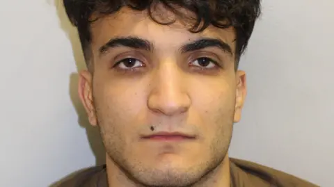 A police mugshot of Sorosh Amini who is looking directly at the camera with long curly black hair which covers his fringe. he has a shaved beard resulting in stubble and is wearing a brown tshirt against a grey background