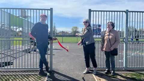 Three people stood outside the multi-games use area. They have just cut a red ribbon and are opening the area to the public. There's a tall man in a blue umper to the left and two women stood to the right.