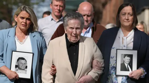 Three women are walking arm in arm outside court toward the camera. Two are holding black and white images of a man in a white and black frame. 
