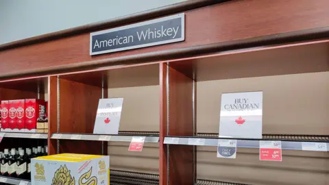 Getty Images An image showing a shelf at a Vancouver liquor store labeled "American Whiskey" that is empty, with signs sitting where the inventory would traditionally be that read BUY CANADIAN. 