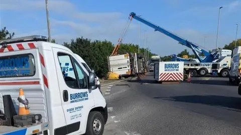 Recovery teams craning the overturned lorry from the side of a roundabout