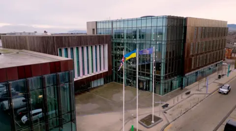 Redcar and Cleveland Council's headquarters photographed from high up. There are three, interlinked square brown buildings. They are clad with the two on either side containing large glass frontages. The middle building, which sits back slightly from the other two, has a large area of wood-effect cladding with windows in the front separated by long, thin white pillars. In front stand three large flagpoles flying the flags of, left to right, the United Kingdom, Ukraine and the council's crest in gold on a blue background.