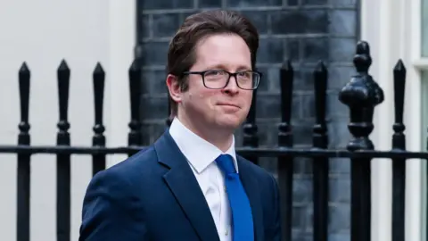 Getty Images Alex Burghart - a man with brown hair, black square framed glasses wearing a navy suit jacket, a white collared shit and a blue tie. He is walking in front of a black iron fence and a black brick building.