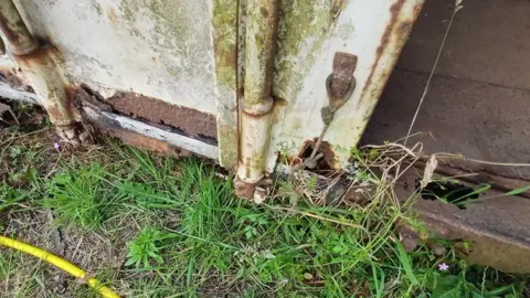 Gloucestershire County Council The base of a very rusty, old shipping container which has holes in it. Patchy, weedy grass is growing on the outside.