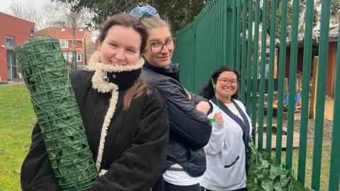 Three young women stand holding a roll of faux shrubbery next to a green metal fence. They are wearing coats and smiling.