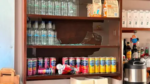 Richard Evans A wooden cabinet with soft drinks on several shelves. The glass window at the front of the cabinet is broken, with broken glass on some of the shelves. 