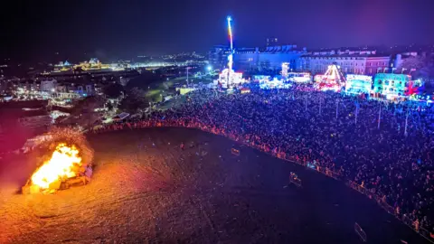 Scott Grenney One Plymouth The photograph shows a large crowd watching the 2023 bonfire. The crowd are held back by barriers and behind them you can see fairground rides.