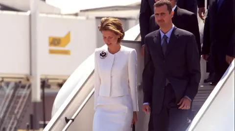 Getty Images Asma al-Assad in white and Bashar al-Assad in black descend a plane staircase.