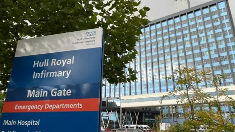 In the foreground, there is a sign that says Hull Royal Infirmary Main Gate and emergency departments. The large oblong hospital building with its blue grey uniformed glass windows stands behind the sign.