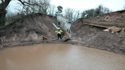 A man in a yellow hi vis jacket and white hard hat stood beside water in a hole after a canal subsided