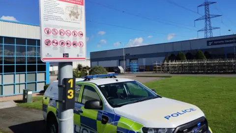 A newly-installed road sign warning of the car cruising rules on a lamppost in front of a police vehicle parked up on a business park.