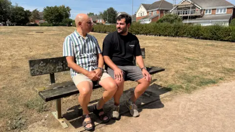 Harjit Singh Nijjer sits on a bench in the park in the sunshine with his son William. Harjit is wearing beige shorts and brown sandals and a green and white striped shirt. He is bald. William is wearing grey shorts and trainers with a black t-shirt. He is sitting with his hands on his knees and has short black hair and a short beard.