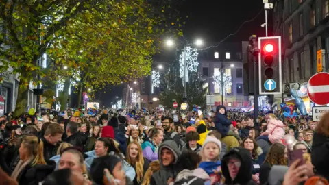 Wolverhampton council Crowds of people in Wolverhampton city centre, with road signs and a traffic light, along with trees in the background and Christmas lights lit up overhead.