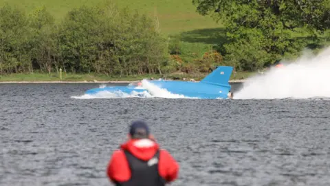Matthew Willis An onlooker in the foreground takes a photograph as Bluebird travels past on Coniston Water. The craft is blue and rocket shaped. There are white bubbly waves around its body and a large white spray from the engine at the rear.