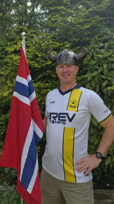 Man in a white and yellow Torquay United football top wears a Viking helmet and holds a red and blue and white Norweigan flag in front of a green hedge 
