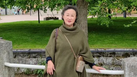 CUH Jia-Ling Ellis is standing by a fence with grass and a tree in the background. She has short, dark hair and is wearing a green shawl-type top.