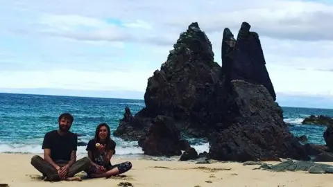 Arthur Serini Arthur and Madi sit cross legged on the beach during their road trip. The sea is behind them, with a dramatic rock jutting out of the waves behind them. 